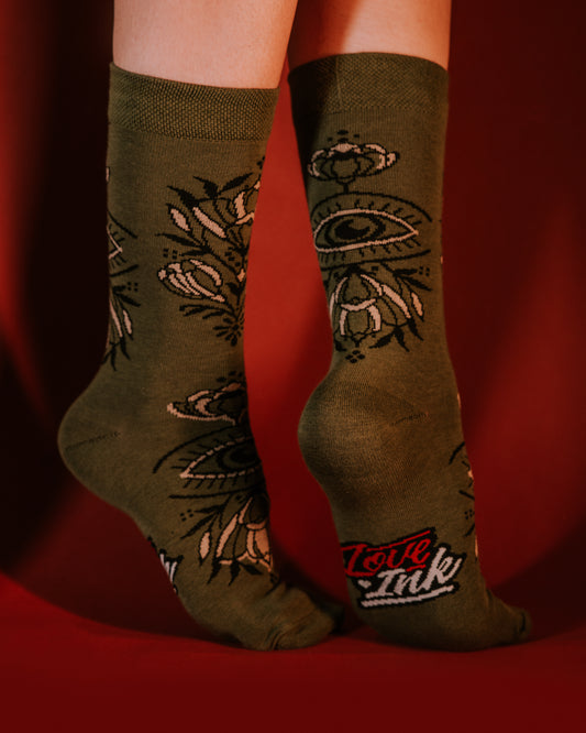Feet wearing green LOVEINK socks with an eye and floral pattern, posed against a red background.