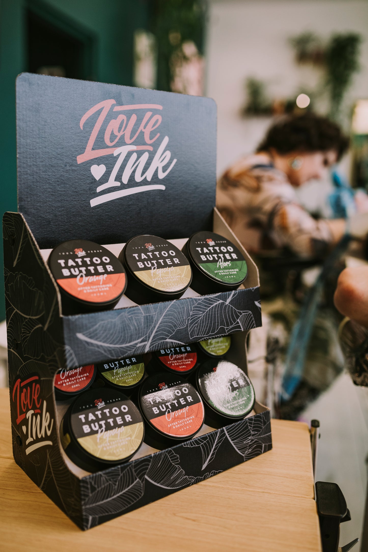 Display stand featuring various Love Ink Tattoo Butter tins in a shop setting. The stand showcases Tattoo Butter in different formulations including Orange, Aloe, and Papaya. In the background, a tattoo artist is seen working on a client in a well-lit, stylish studio.