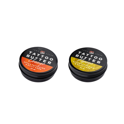 Two tins of Love Ink Tattoo Butter on a transparent background. One tin is labeled 'Tattoo Butter Orange' with an orange design, and the other is labeled 'Tattoo Butter Papaya' with a yellow design. Both tins are 50 ml and are intended for after tattooing and daily care.