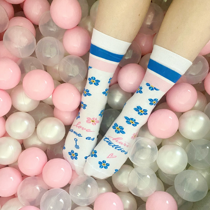 Feet wearing white socks with blue and yellow flower patterns, blue stripes at the top, and the Love Ink logo, lying in a ball pit filled with pink and white balls.