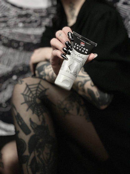 Close-up of a person with tattooed arms and legs holding a tube of Tattoo Cream by LoveInk, emphasizing its use for tattoo care.