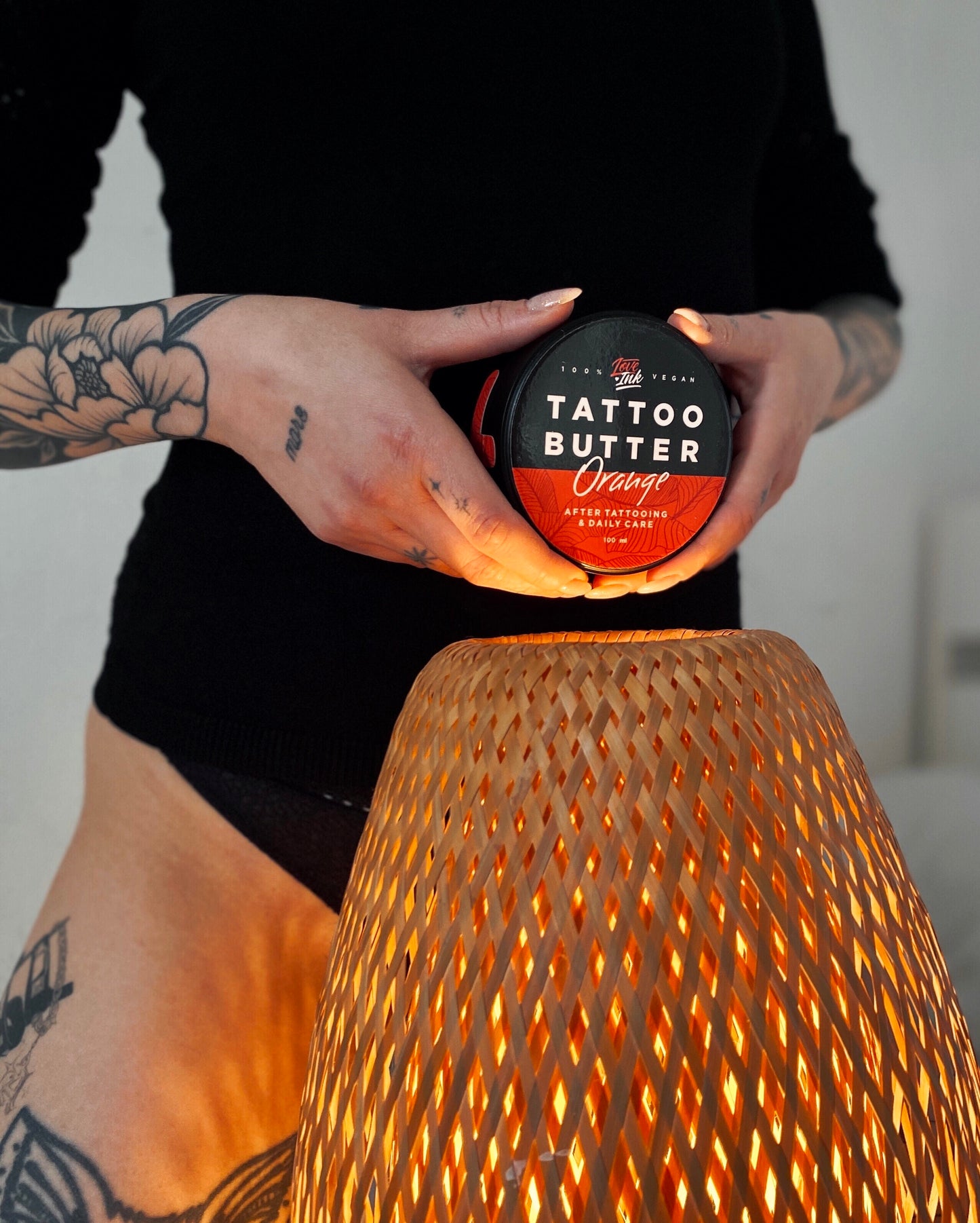 A person holding a jar of Tattoo Butter Orange by LoveInk near a woven lamp emitting a warm glow. The scene emphasizes the soothing and daily care benefits of the product, 100ml.