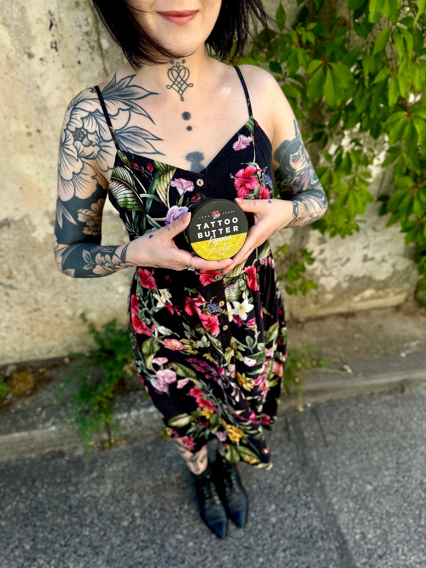 Person in a floral dress holding a jar of Tattoo Butter Papaya by LoveInk. The background features a natural setting with greenery.