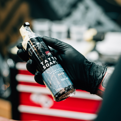 Hand in a black glove holding LoveInk's Tattoo Soap Silver bottle, ready for use during the tattooing process. Red tool cabinet in the background.