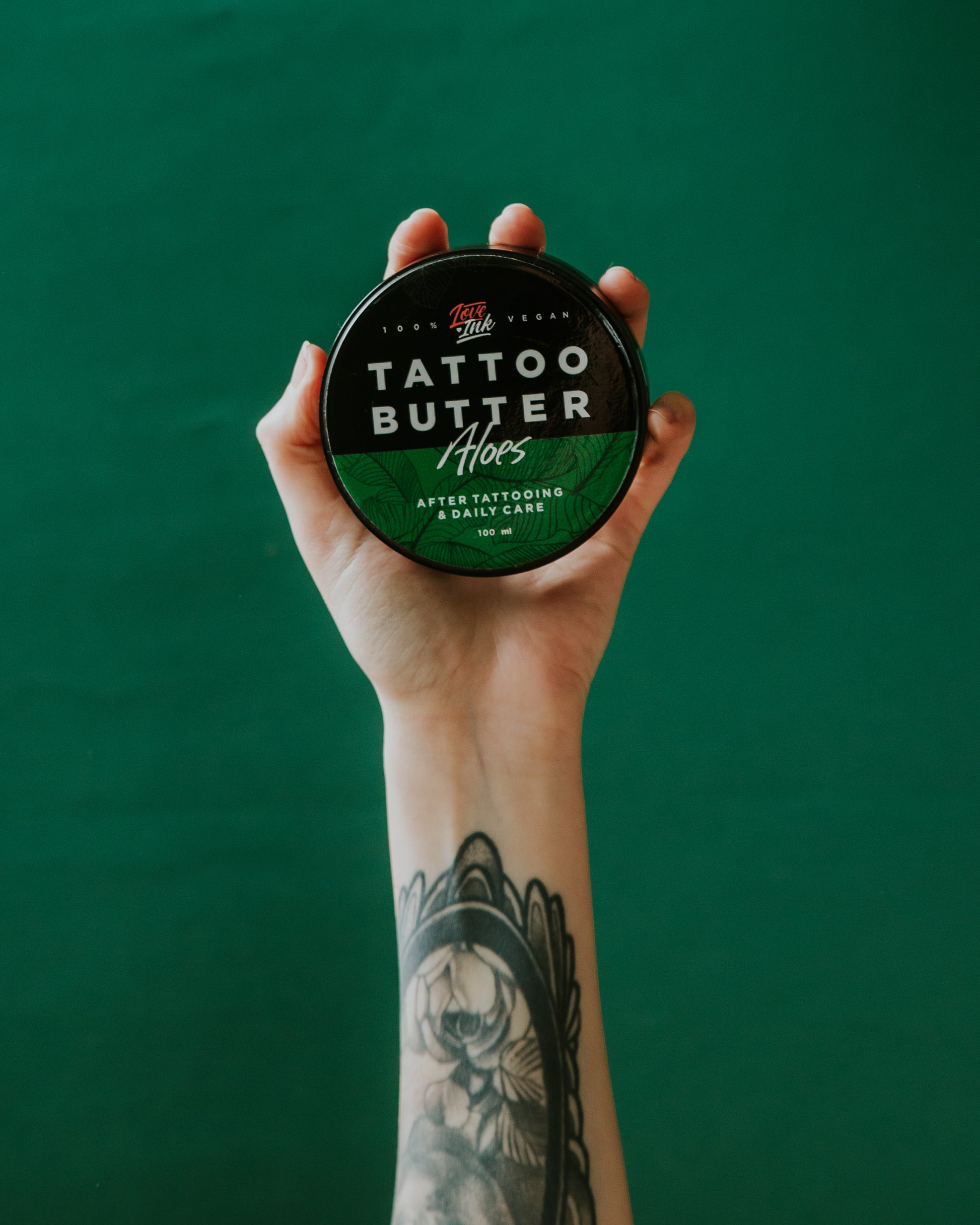 A hand holding a tin of Love Ink Tattoo Butter Aloe against a green background, with a floral tattoo visible on the wrist.